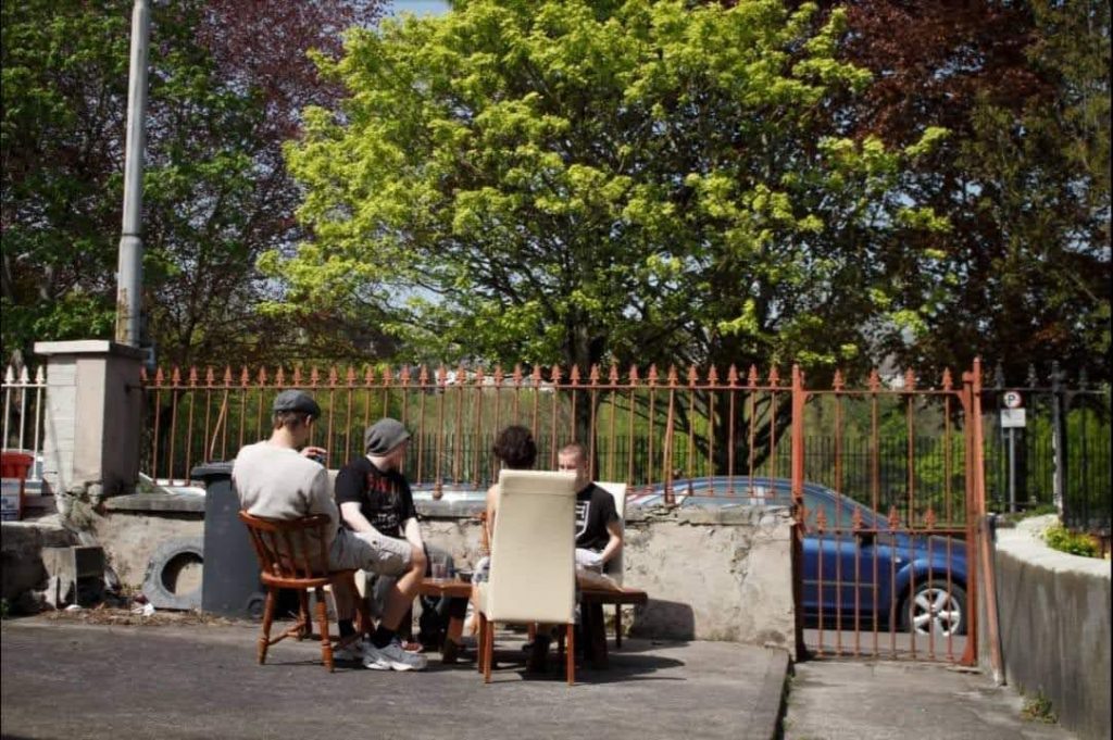 Four members sit at a table brought out to the front yard. The leaves of the trees in the background paint the sky in bright and vibrant greens and autumnal reds