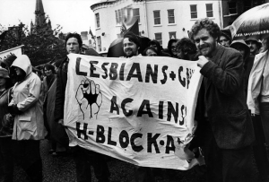 Three men hold up a banner that reads "LESBIANS + GAYS AGAINST H-BLOCK-A"